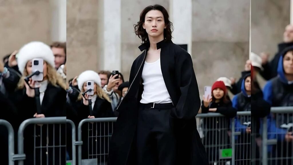 Sang Heon Lee in a black coat and white shirt walks confidently down a city street.