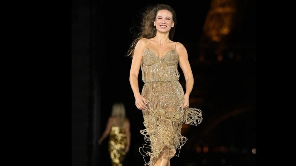 Serenay Sarikaya in a shimmering gold dress confidently walks down the runway during a fashion show.