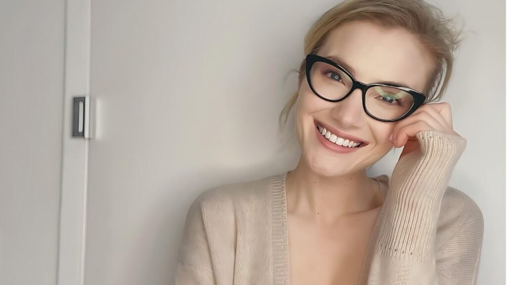 Skyler Samuels with black glasses, leaning against a pale wall, hand resting on her cheek, wearing a beige cardigan.