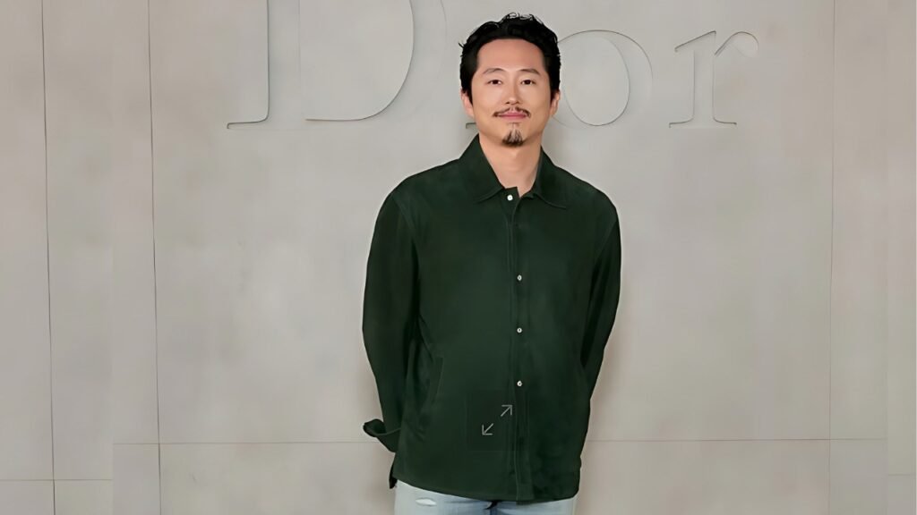 Steven Yeun wearing a green shirt and jeans stands in front of a prominent Dior logo.