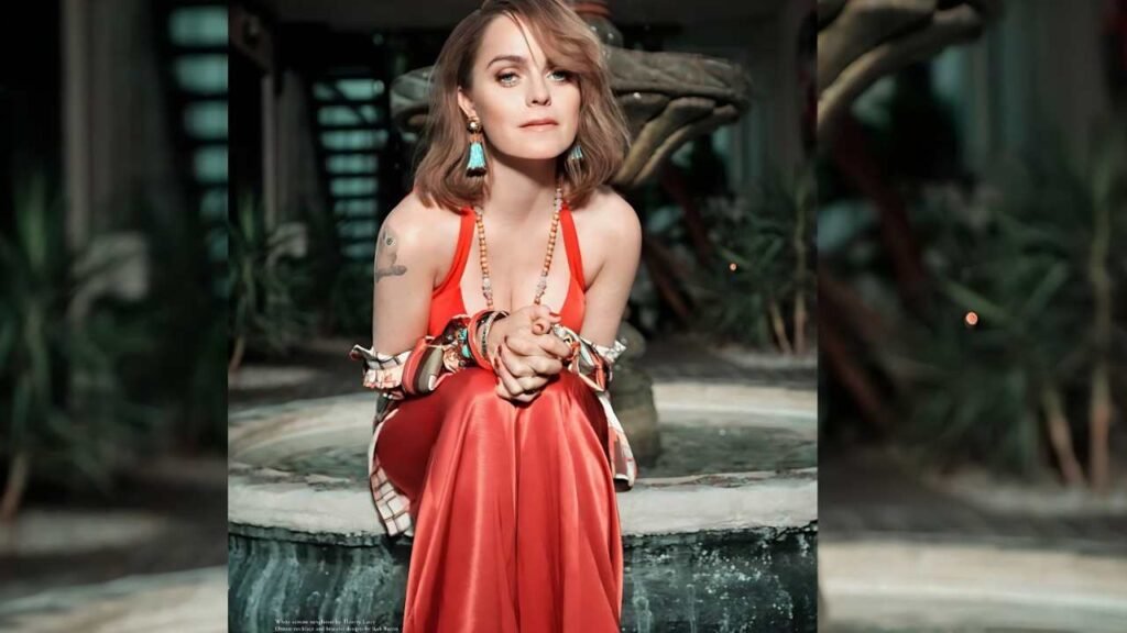 Taryn Manning sits on a stone fountain edge wearing a red satin dress, turquoise earrings, bracelets, hands clasped, garden backdrop.