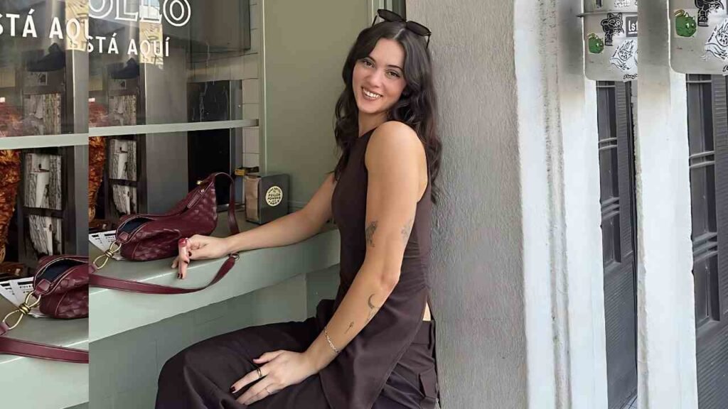Tatum Dahl in a dark brown sleeveless dress sits at a counter outside, sunglasses on her head, maroon handbags nearby.
