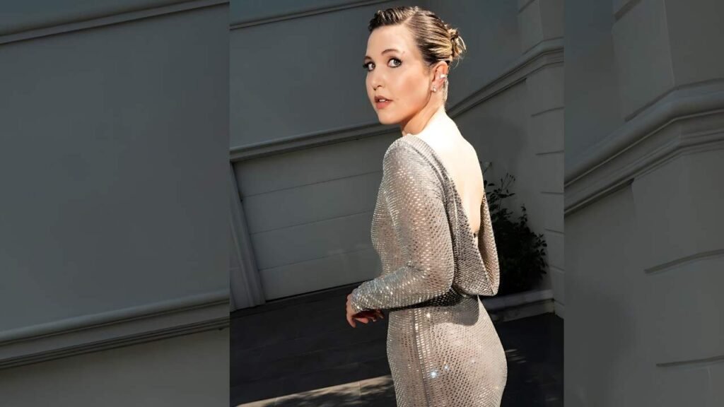 Taylor Tomlinson in a silver sequined gown with a low back, hair in an updo, glancing over her shoulder beside a pale building.