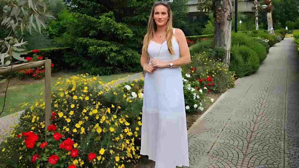 Timea Babos in a white dress stands on a sidewalk, surrounded by greenery and urban scenery.
