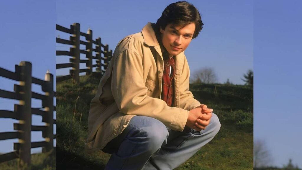 Tom Welling wearing a jacket and jeans kneels on the grass, appearing contemplative in a natural outdoor setting.