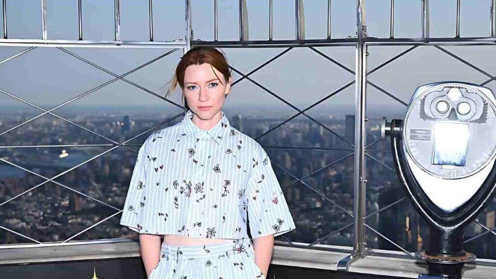 Valorie Curry in a blue shirt and pants stands on a chair, reaching for an item above her.