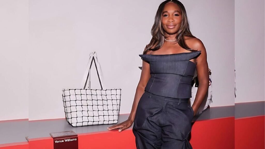 Venus Williams in a black top and pants stands beside a white bag, smiling confidently at the camera.