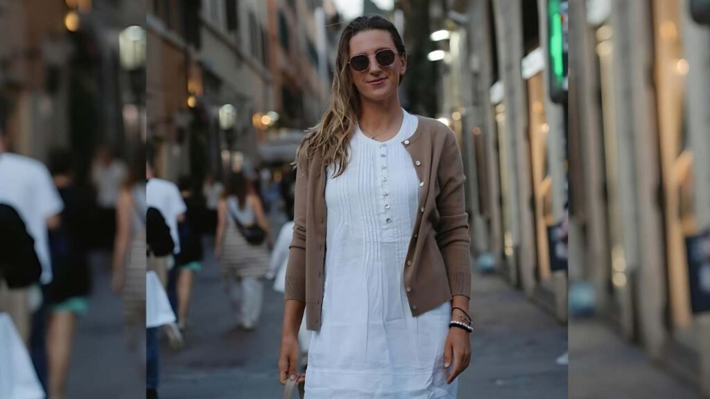 Victoria Azarenka wearing a white dress and a brown cardigan stands confidently, showcasing her stylish outfit.