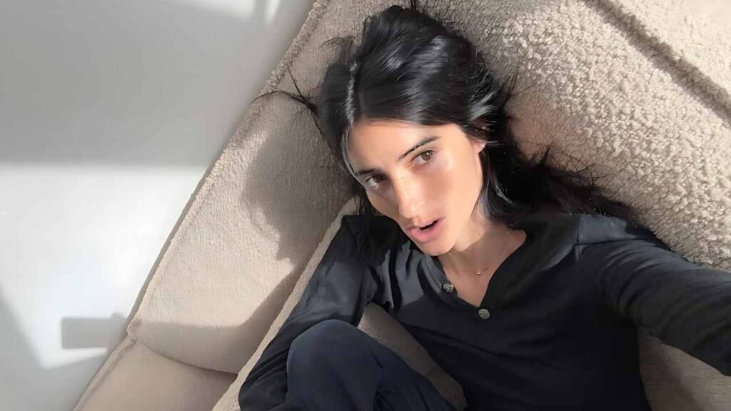 Virgo Martinez with black hair wearing a dark shirt lounges on a plush beige couch, bathed in sunlight, looking to the camera.