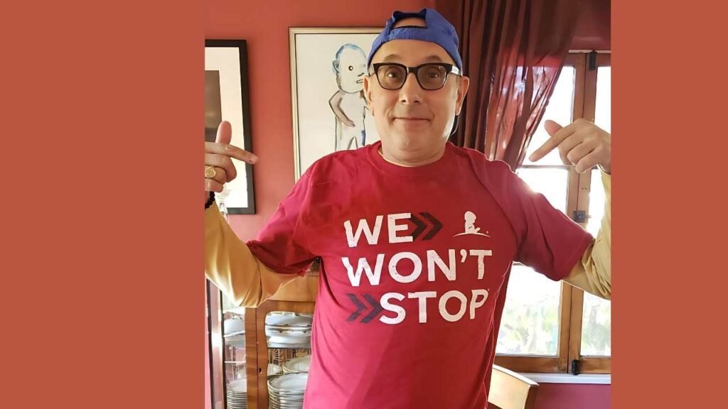 Willie Garson wearing a bright red shirt featuring the slogan "We Won't Stop," exuding a sense of motivation and resilience.