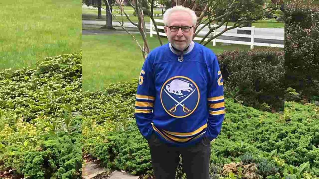 Wolf Blitzer with glasses stands in a green garden wearing a blue Buffalo Sabres hockey jersey with a crossed-sabres logo.