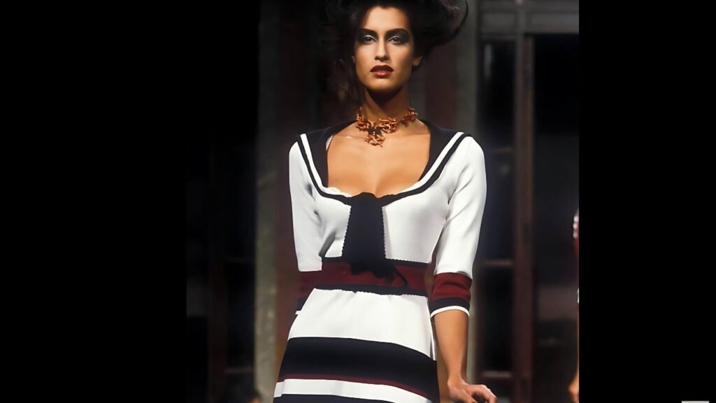 Yasmeen Ghauri on a runway wears a white sailor-top with black trim, a black tie, maroon waist bands, and bronze necklace.
