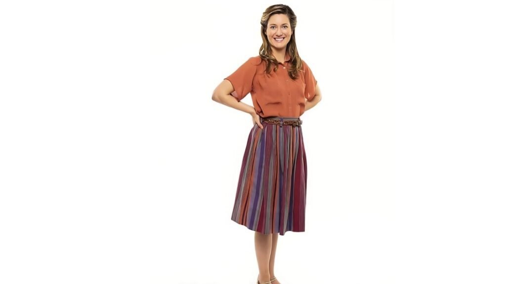 Zoe Perry wearing a skirt and an orange top poses confidently for a photograph.