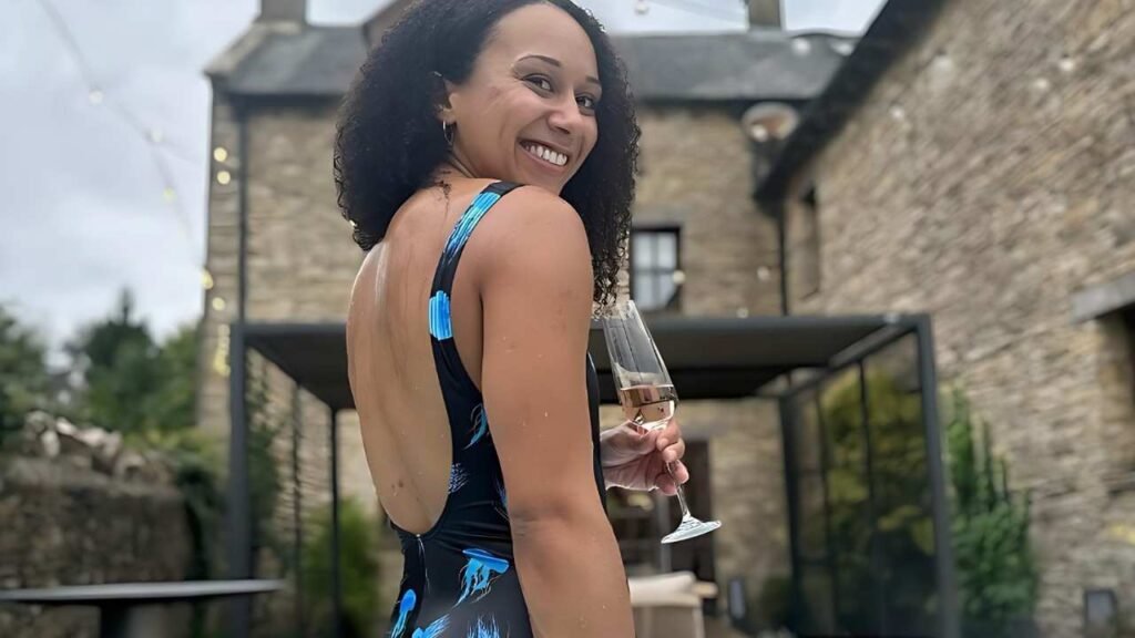 Zoe Smith in a blue backless swimsuit with blue accents turns toward the camera, holding a champagne flute in a stone courtyard.