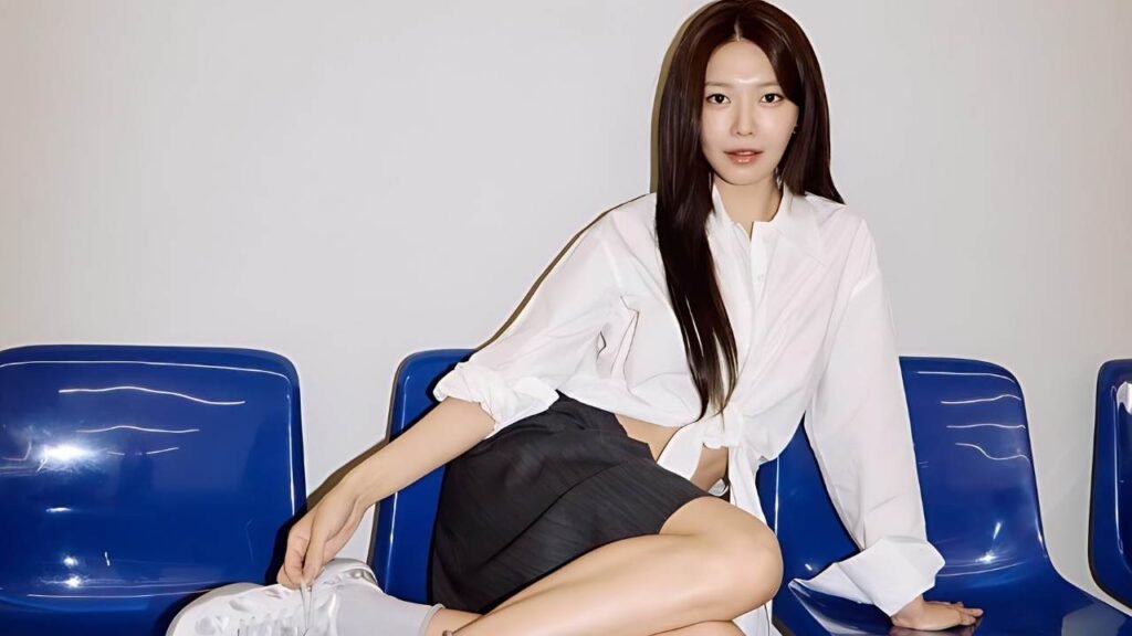 Choi Soo-young sits on a blue chair with her legs crossed, wearing casual attire and smiling at the camera.